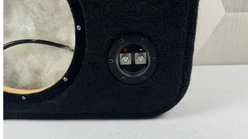Close-up of a black car audio enclosure with wiring and connectors on a white surface.